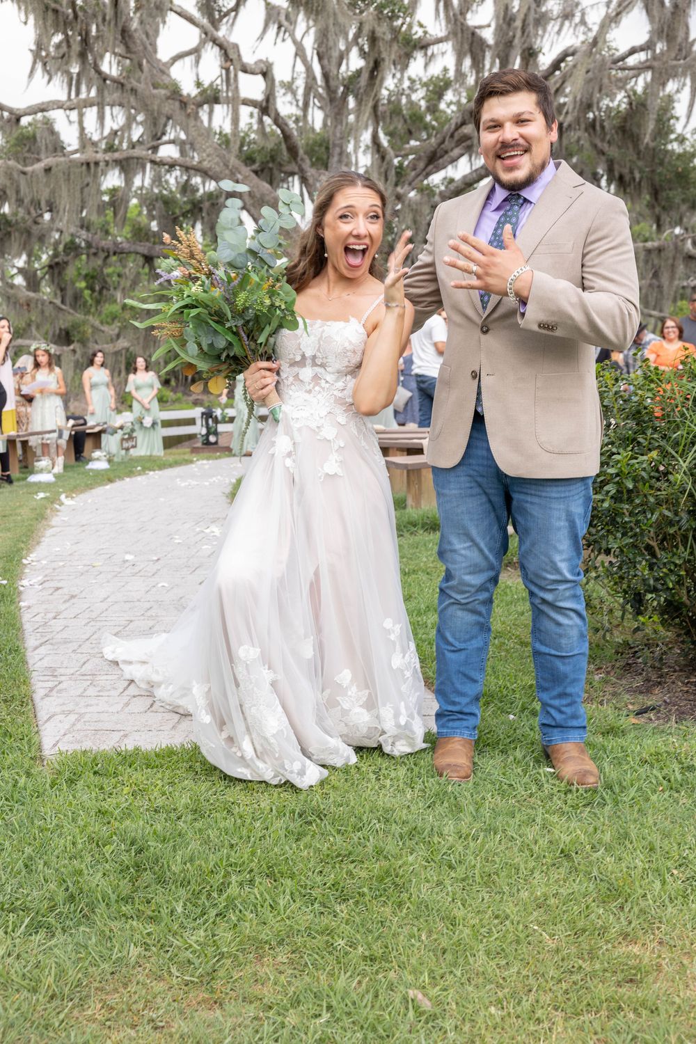 Joyful newlywed couple celebrating after outdoor wedding ceremony, bride in floral gown holding bouquet, groom in tan blazer.