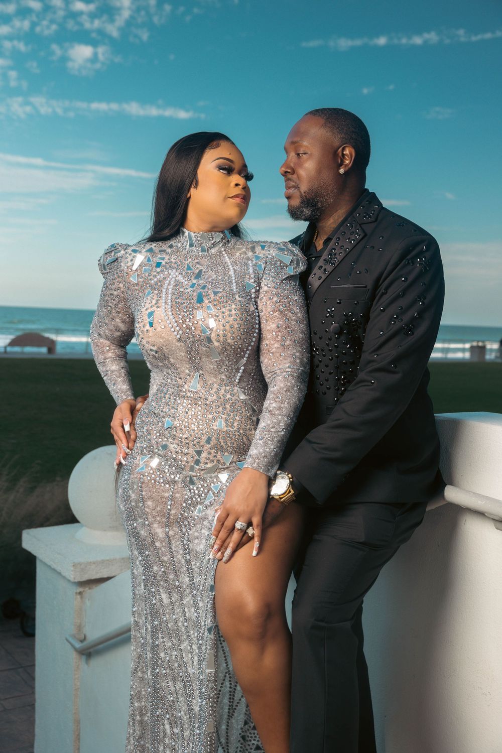 Elegant couple posing oceanside, woman in silver sequin gown, man in black embellished suit, blue sky backdrop.