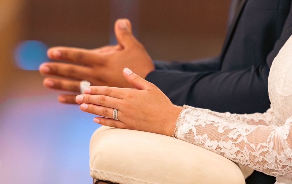 Close-up of bride and groom hands clasped together, showing wedding rings and lace bridal gown.