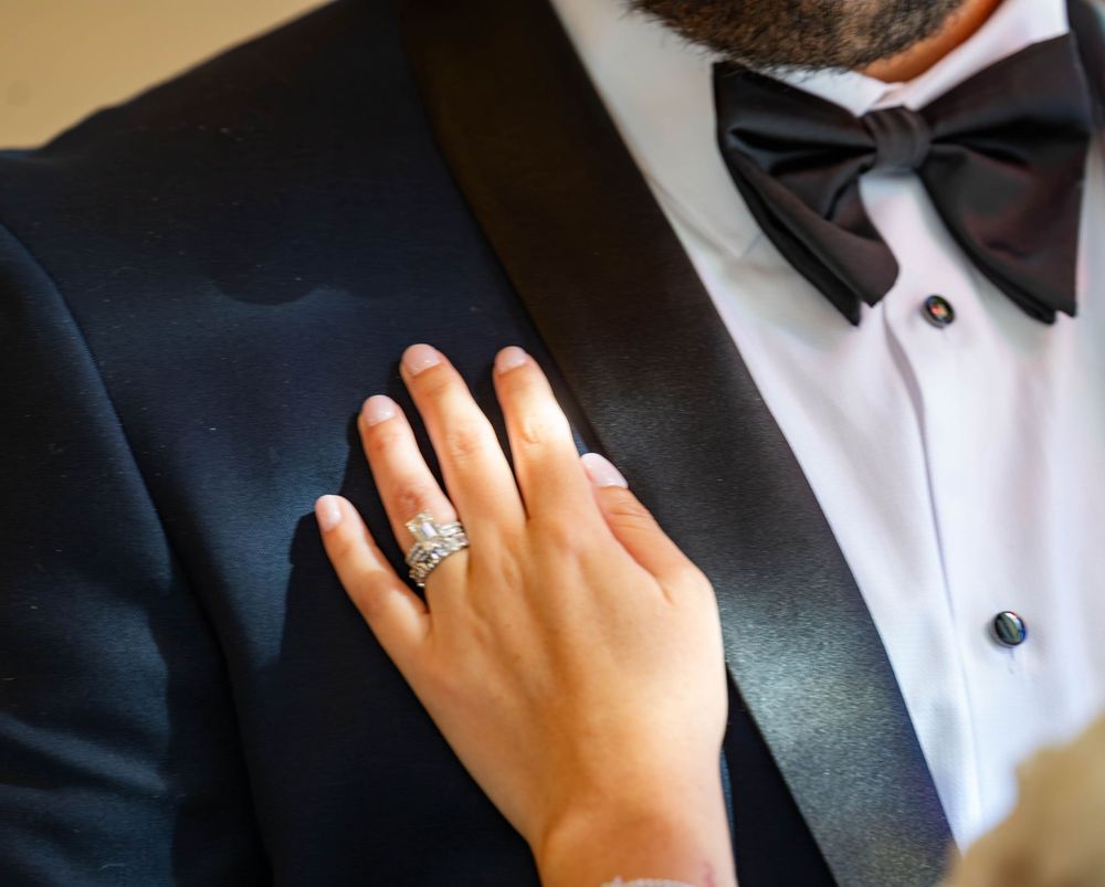 Bride's hand with diamond ring rests on groom's navy tuxedo jacket with black bow tie.