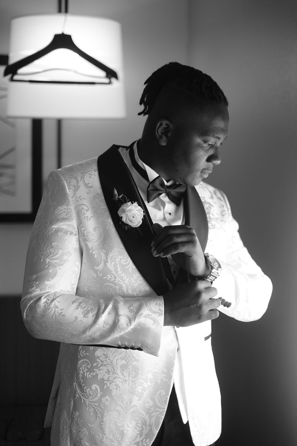 Black and white photo of a groom in a white floral tuxedo adjusting his cufflinks before his wedding ceremony.