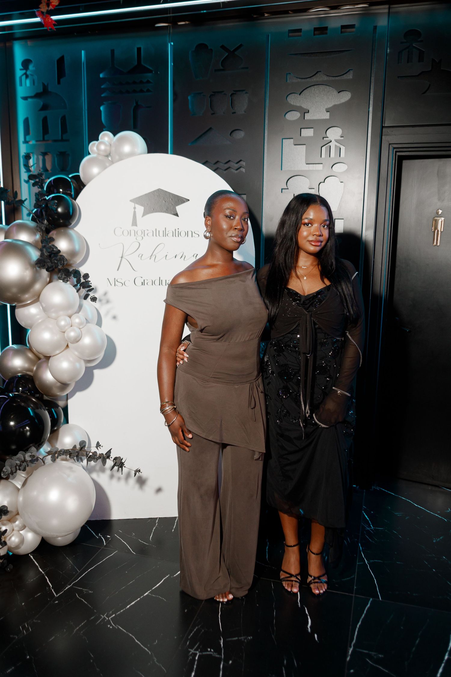 Two women posing at a graduation celebration party with black and silver balloon arch backdrop.
