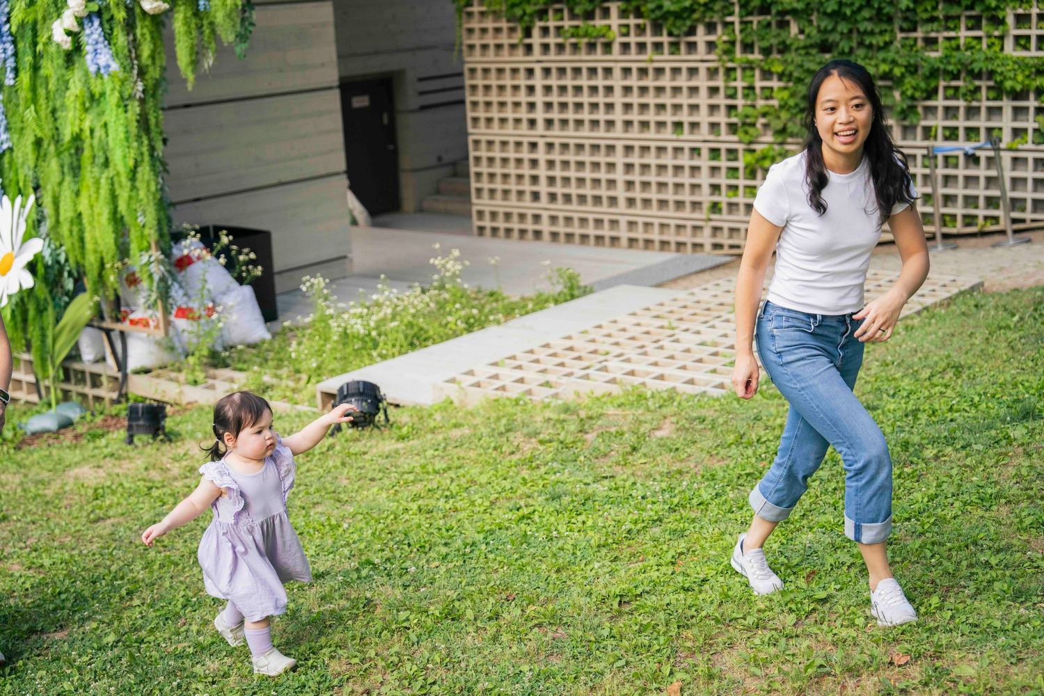 A mother and young daughter playing together on green grass in a sunny backyard garden setting.