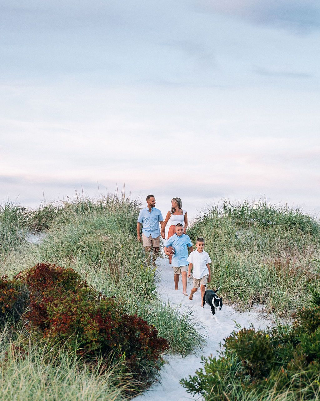 Family with two boys and a dog walking along a sandy beach path through coastal dunes at sunset.