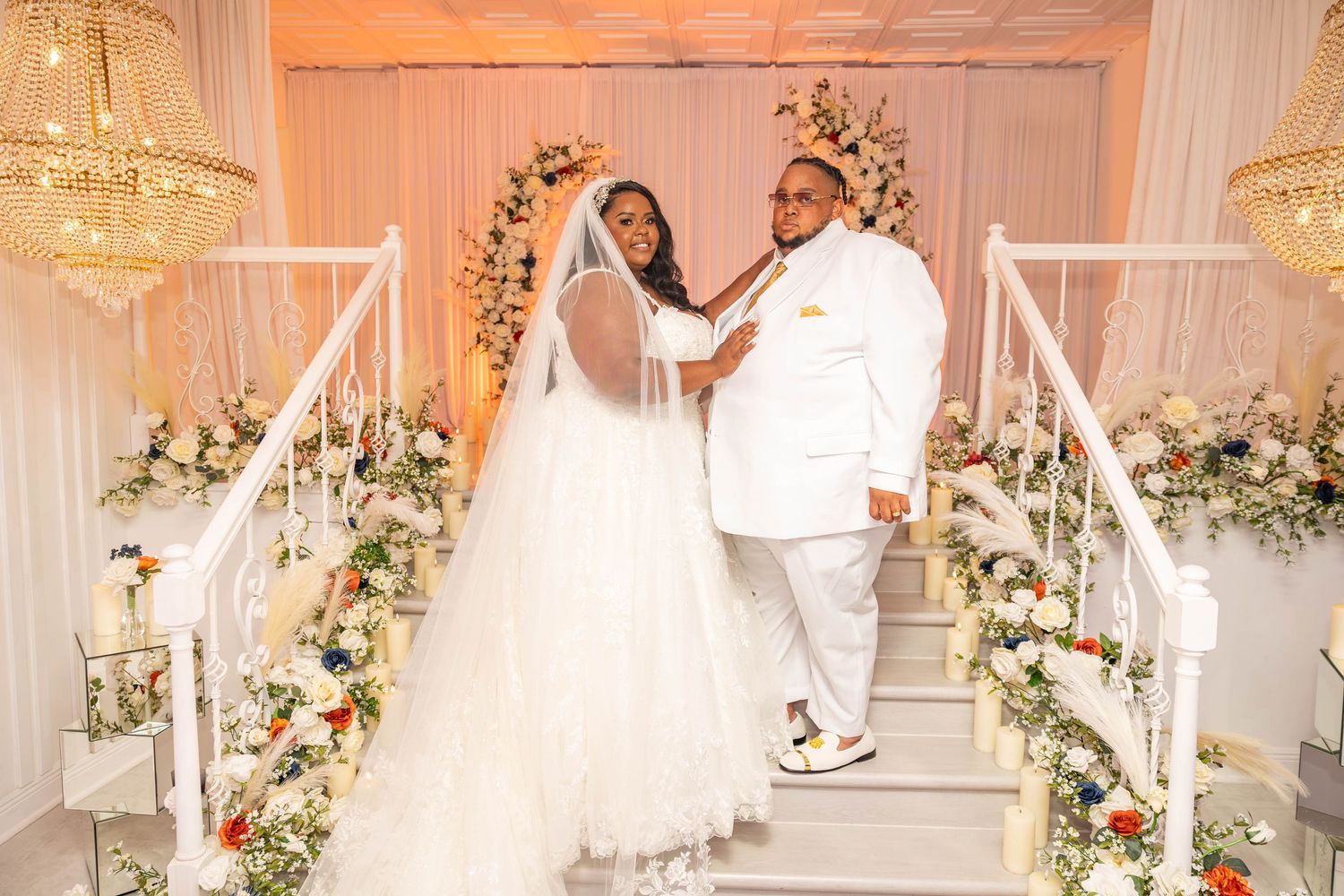Bride and groom pose on white staircase adorned with flowers at elegant wedding ceremony.