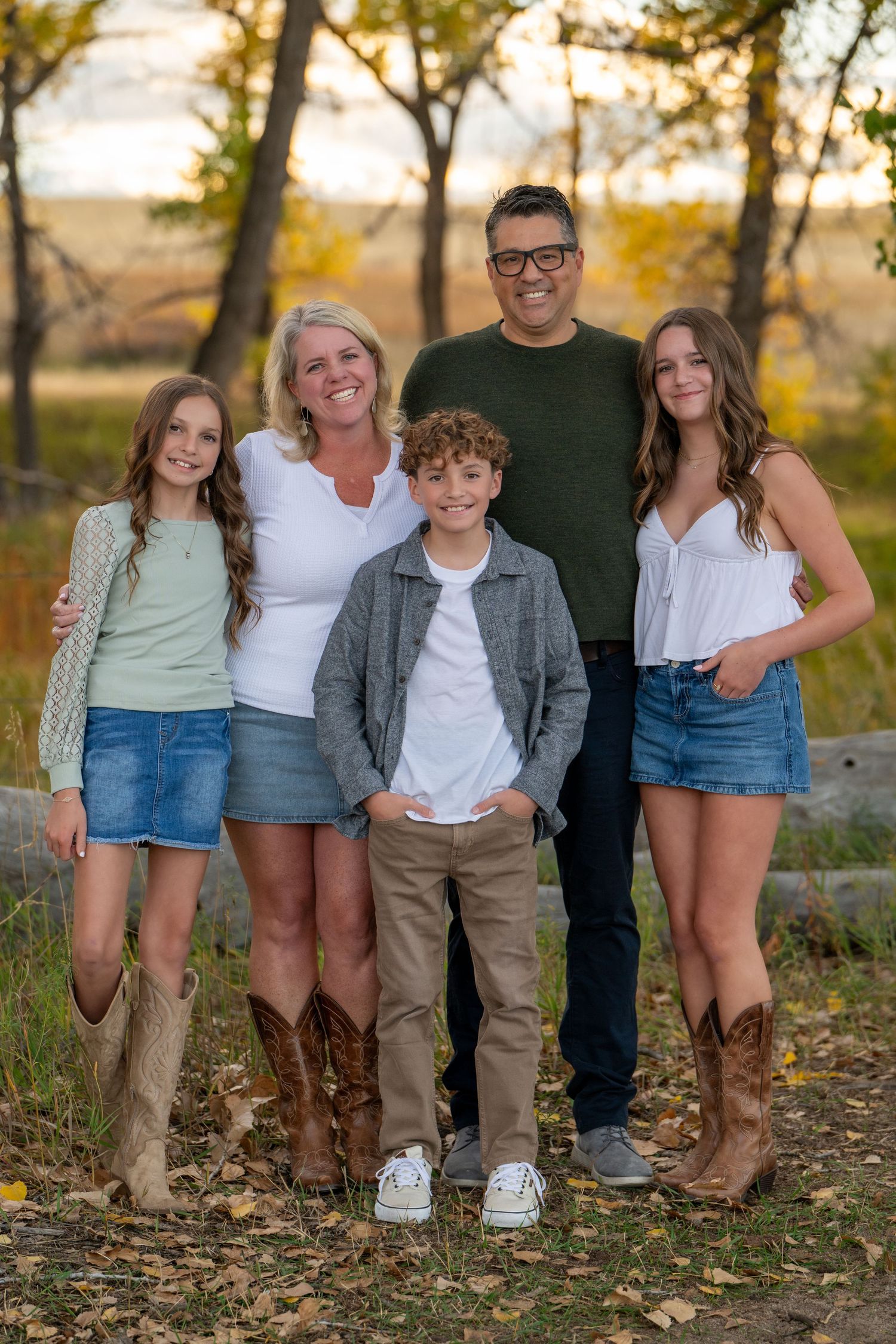 Family of five smiling outdoors in autumn, wearing casual western attire including denim skirts and cowboy boots.