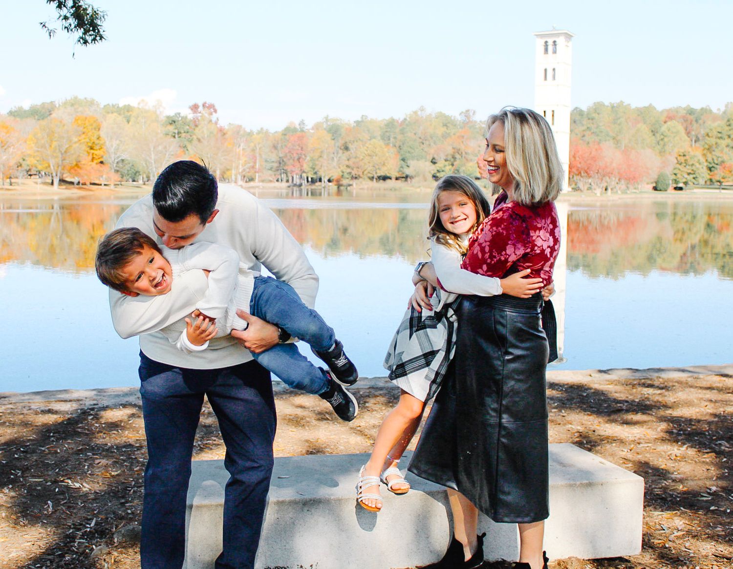 Family of four laughing and playing by a lake with autumn foliage in the background.