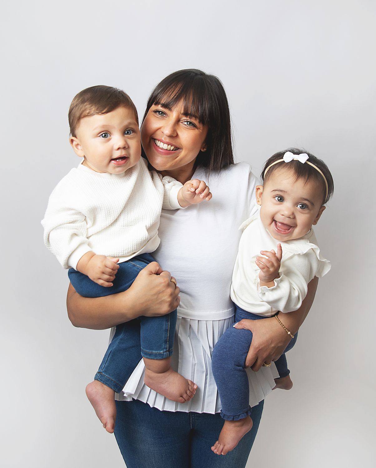 A smiling woman holds two happy babies dressed in white outfits against a white studio background. During a family photoshoot in Bexley