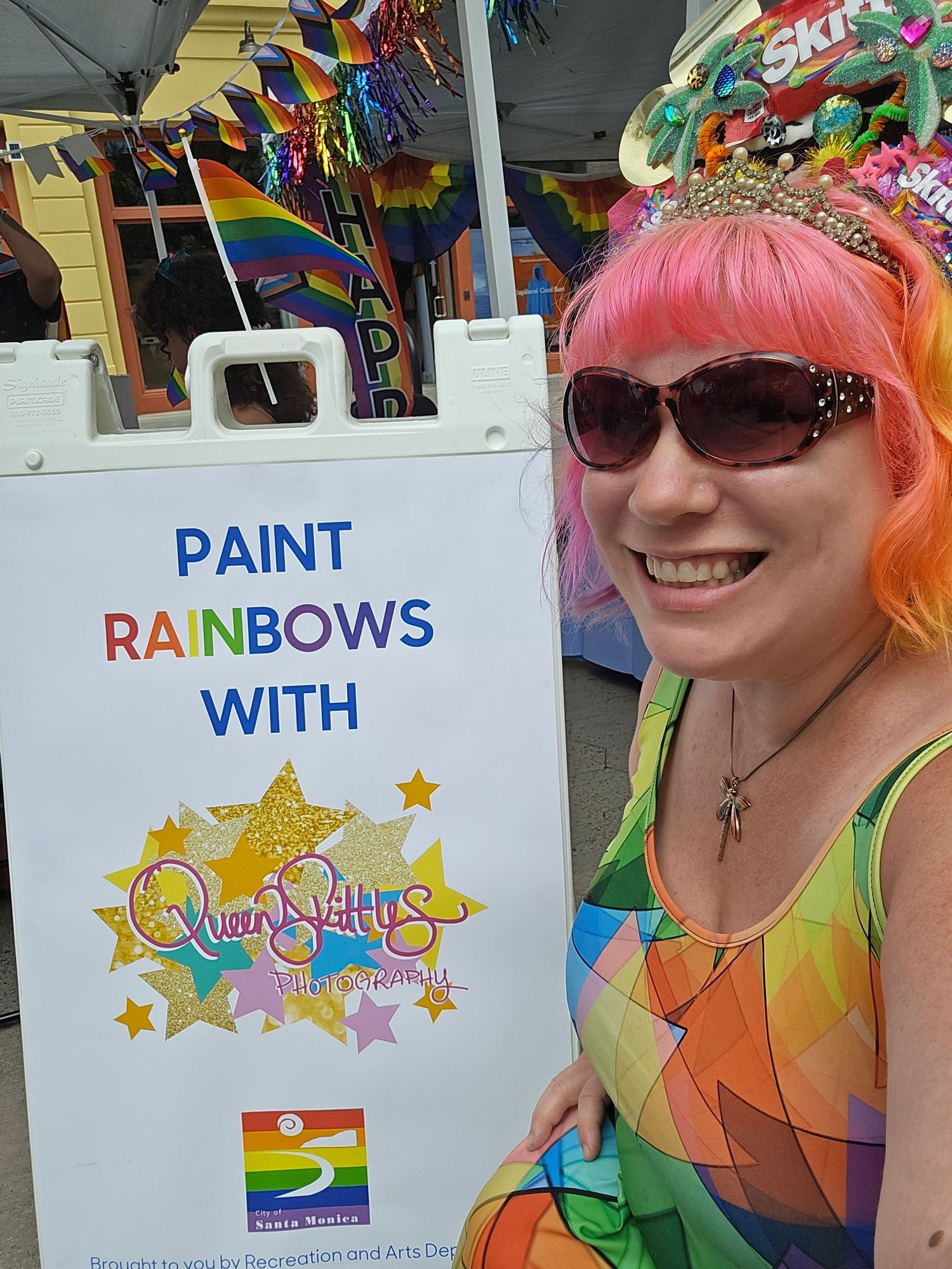Woman with pink hair and rainbow outfit smiles beside a Paint Rainbows With Queen Bettys sign at Pride event.