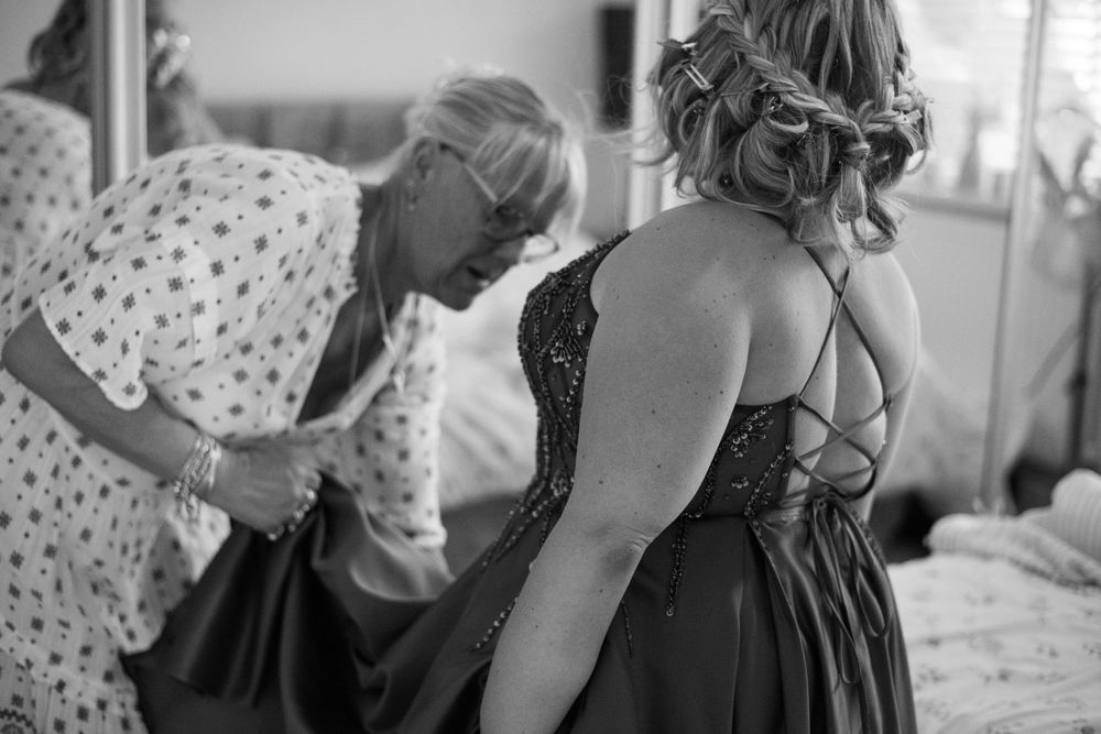 Bridesmaid in lace-back dark gown being helped with dress by woman in floral top, black and white wedding photo.