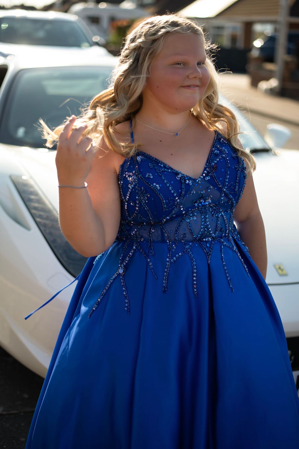 Young woman in royal blue sequined ball gown poses outdoors near white sports car for prom.