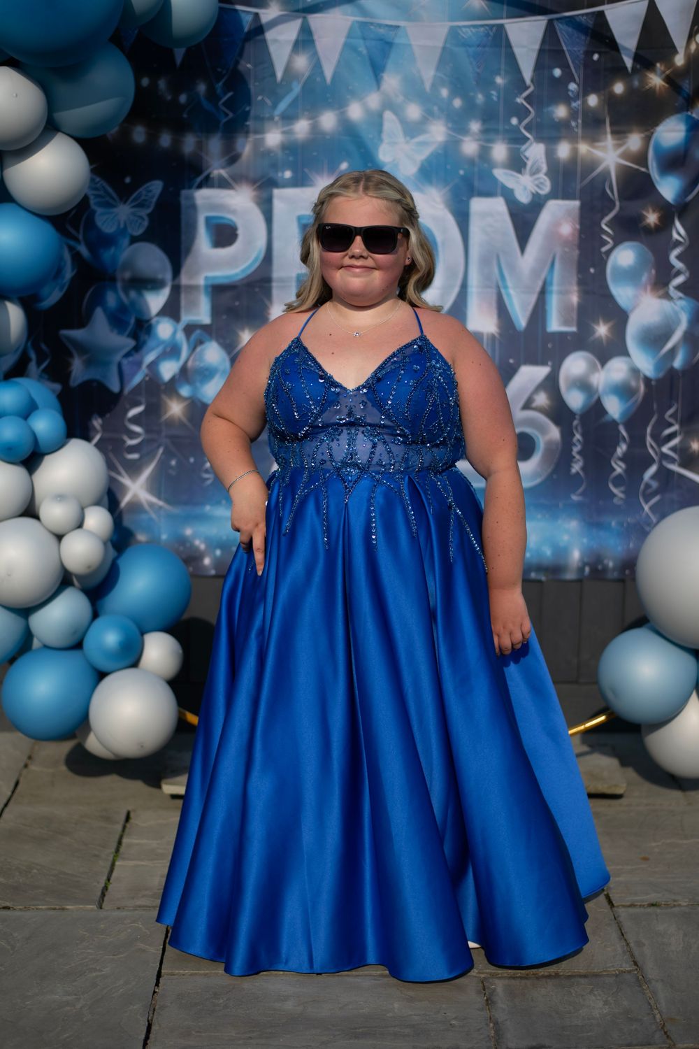 Teen girl in royal blue beaded prom dress with sunglasses posing in front of blue balloon prom backdrop.