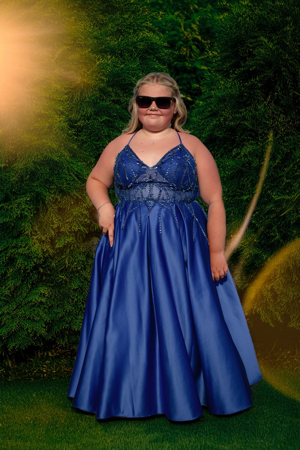 Young woman in royal blue beaded satin prom gown with sunglasses posing outdoors against green hedges.