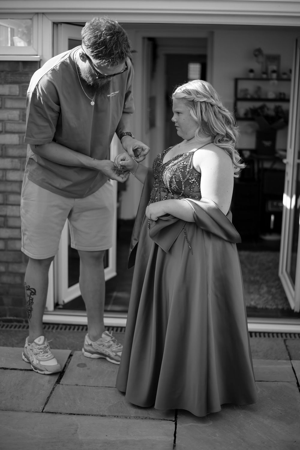 Man helping young woman in elegant gray prom gown outside home in black and white photo.
