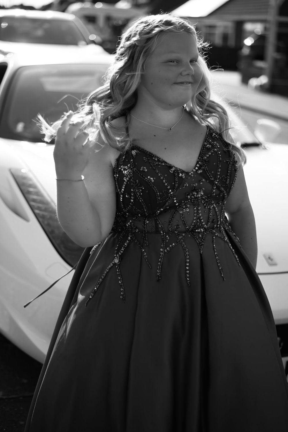 Young woman in sequined formal gown poses beside a white sports car in black and white photo.