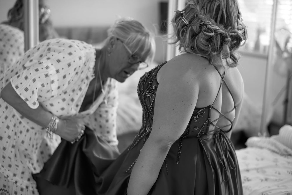 Black and white photo of a woman having her lace-back formal gown fastened by another woman before a wedding.
