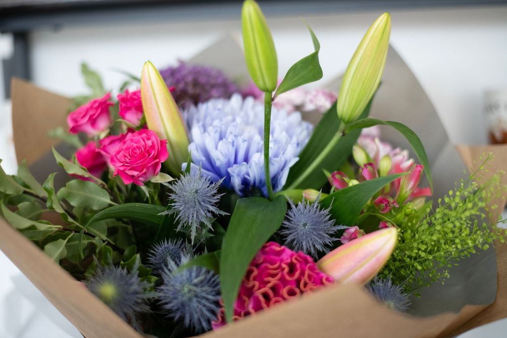 Vibrant mixed bouquet featuring pink roses, blue hyacinths, white lilies, and thistle wrapped in brown kraft paper.