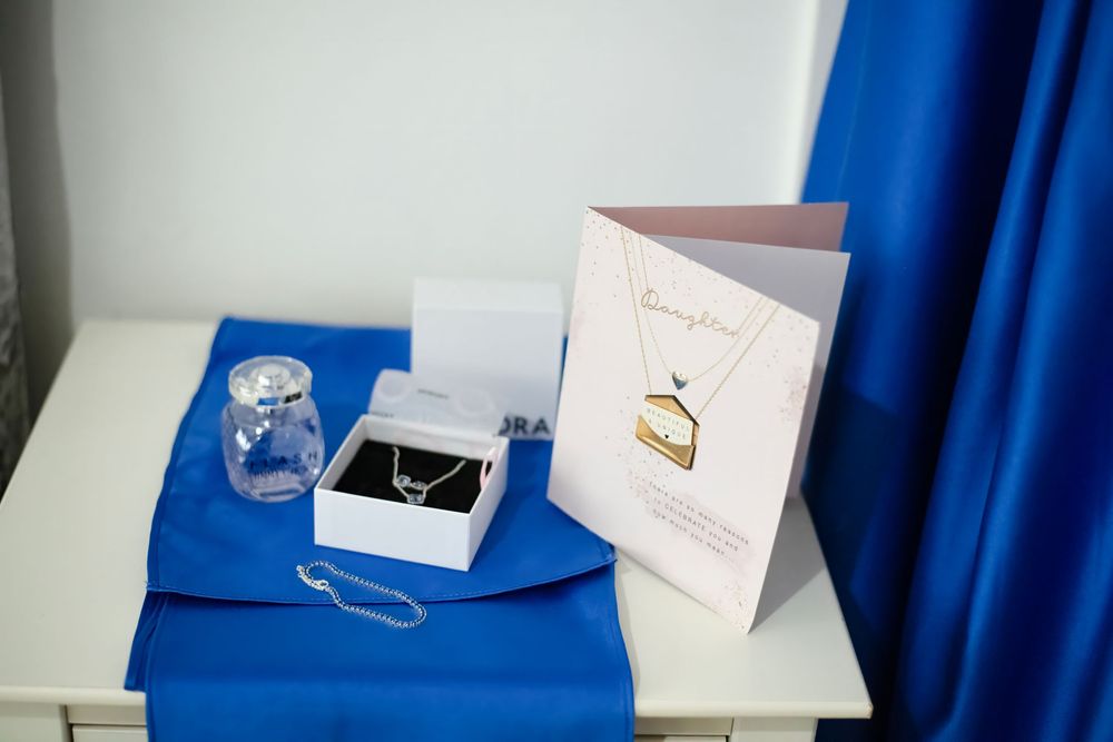 Elegant jewelry gift set with necklace in white box, greeting card, and blue fabric on white table.