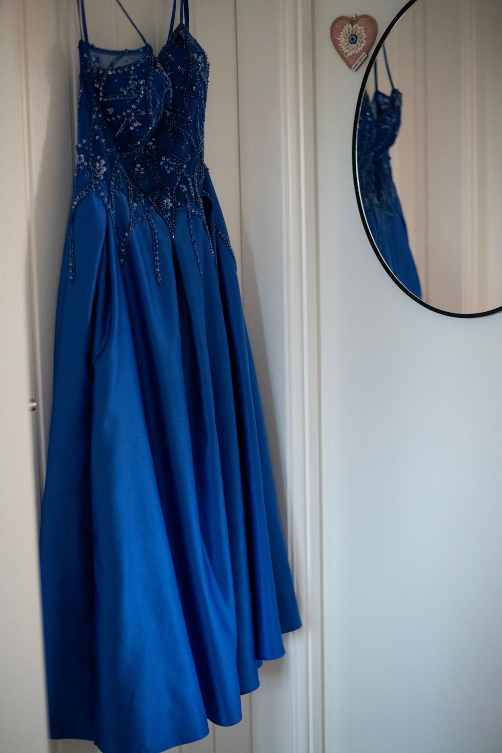 Royal blue satin prom dress with beaded lace bodice hanging on white door beside round mirror.