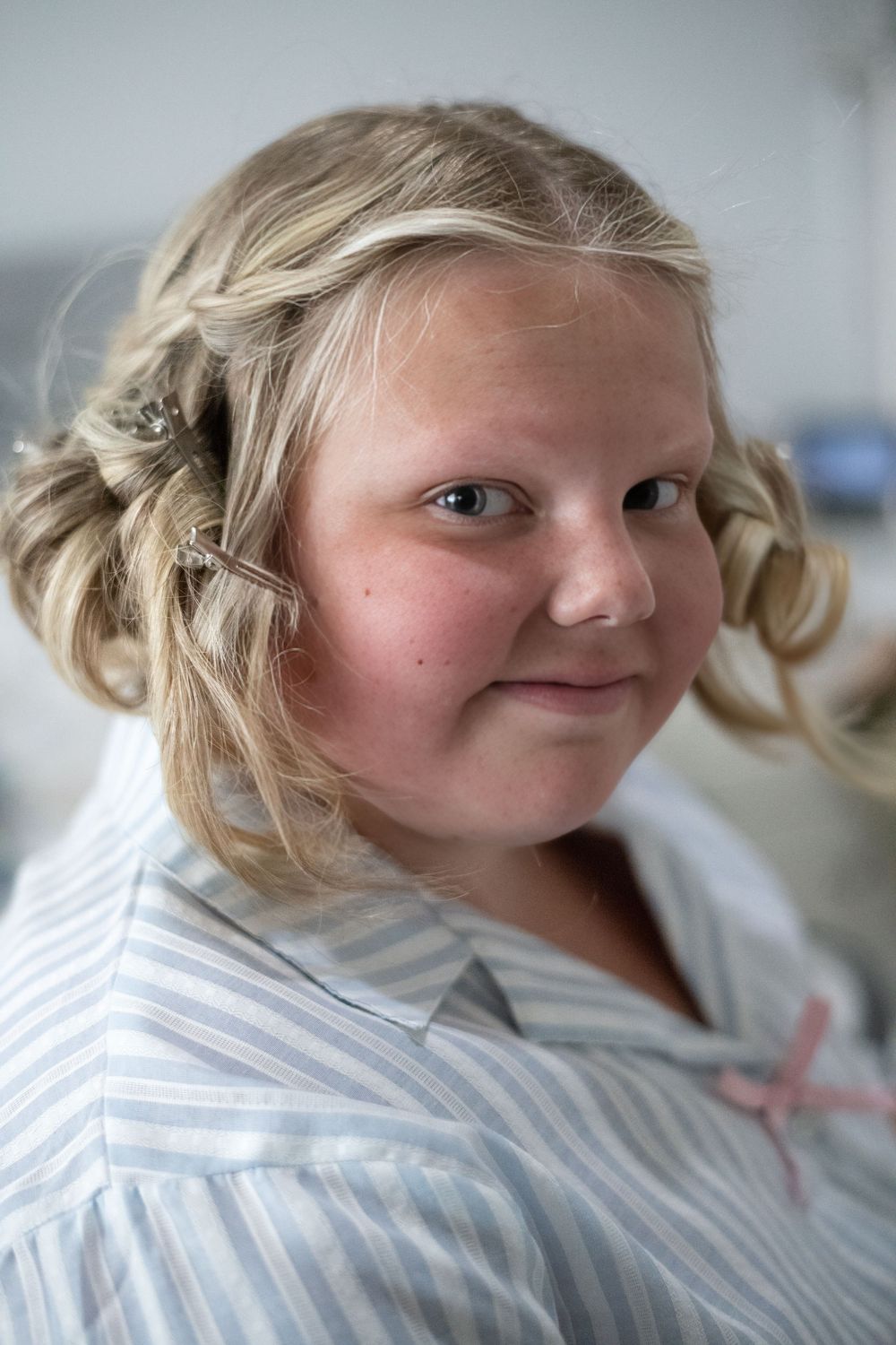 Young blonde girl with curly hair wearing a striped blue and white shirt with a pink bow, smiling softly.