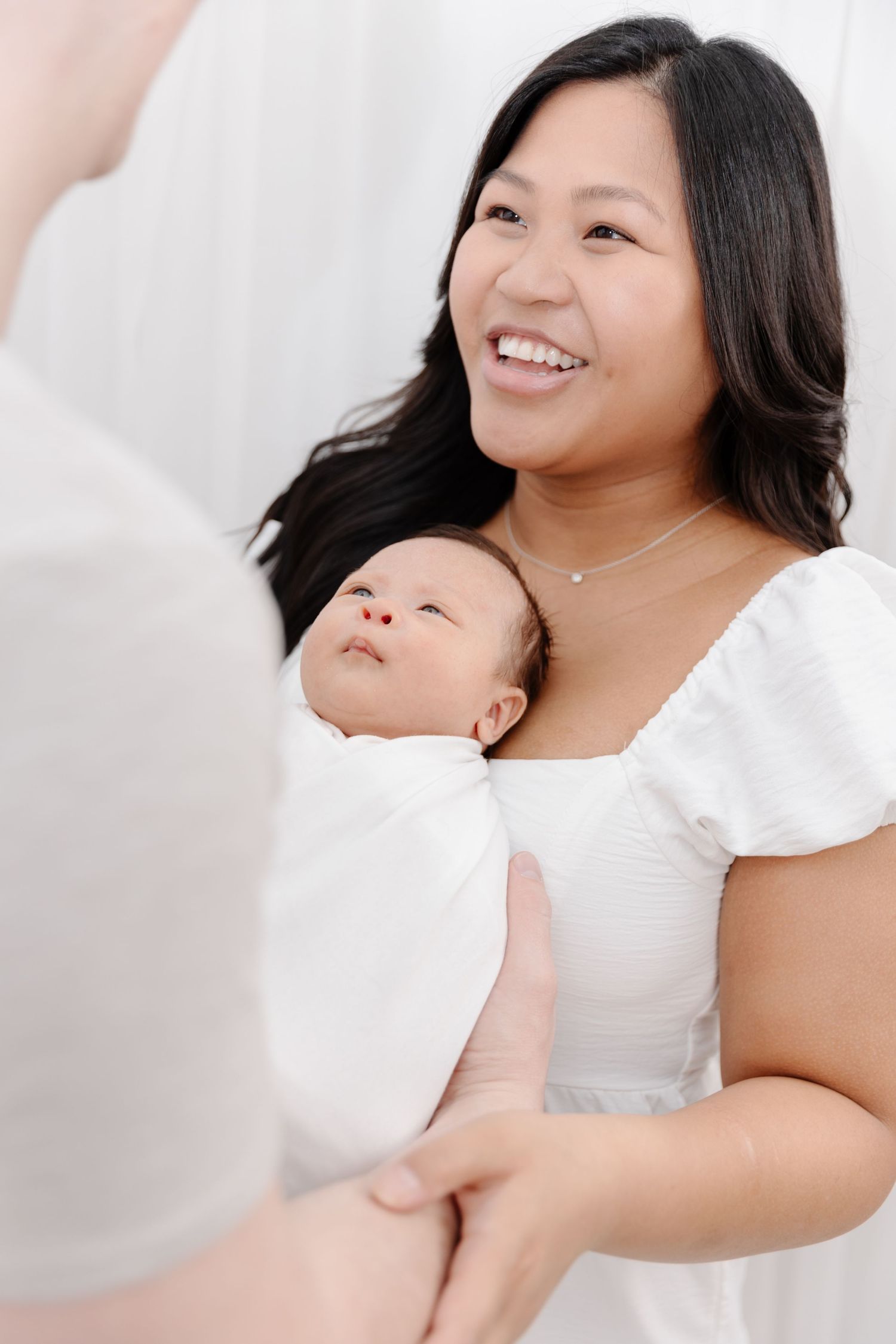 Smiling Asian mother in white dress holds swaddled newborn baby while looking up at partner in bright studio setting.