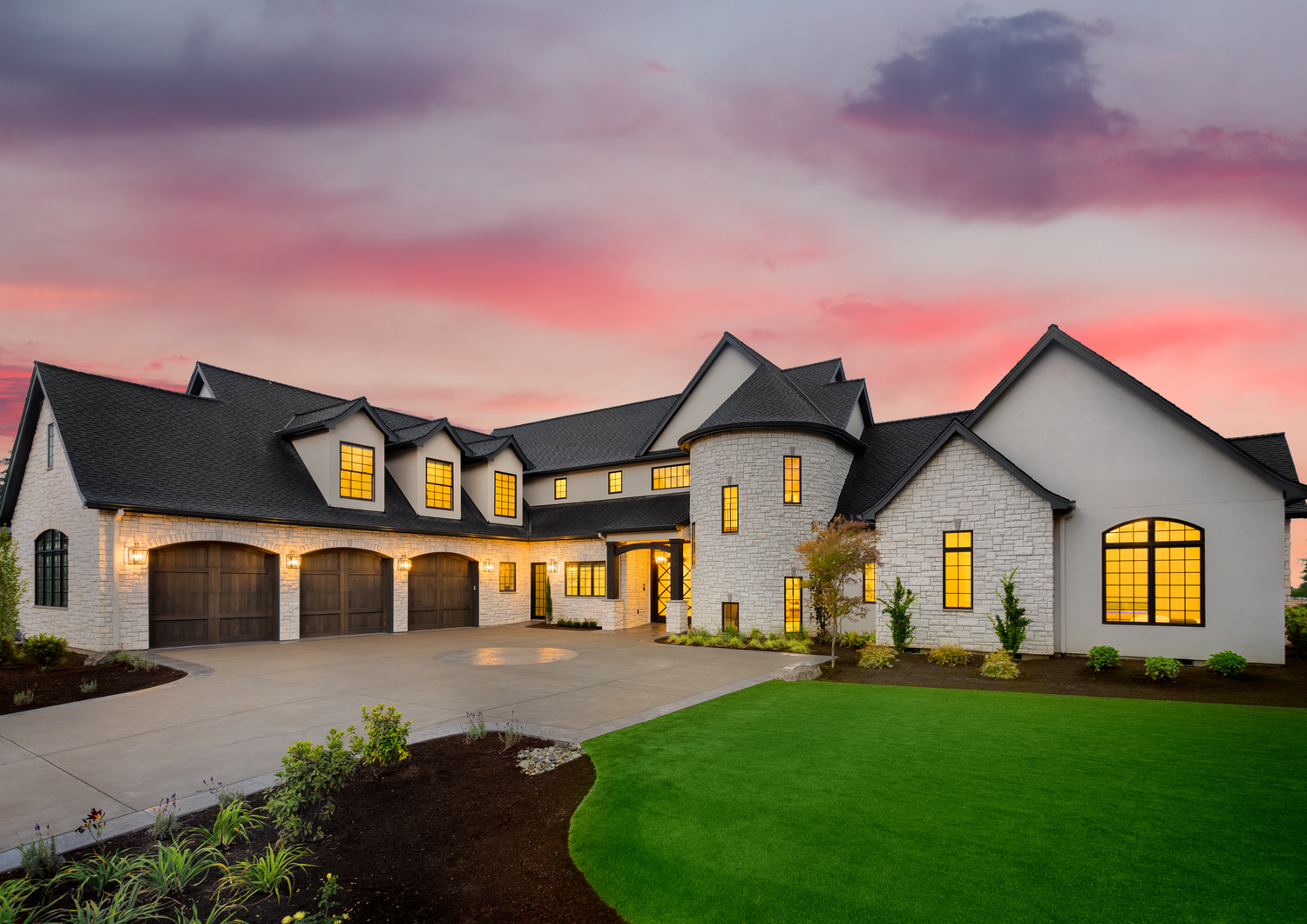 Luxury modern farmhouse exterior at sunset with white stone facade, dark roof, triple garage, and lush green lawn.
