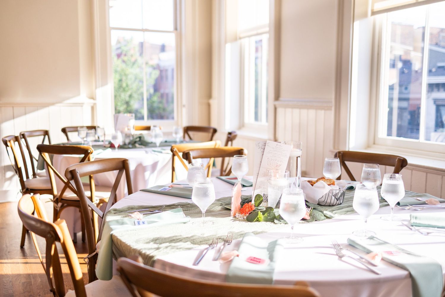 Elegant round dining tables with floral tablecloths, crystal glasses, and wooden chairs in a bright, sunlit space.