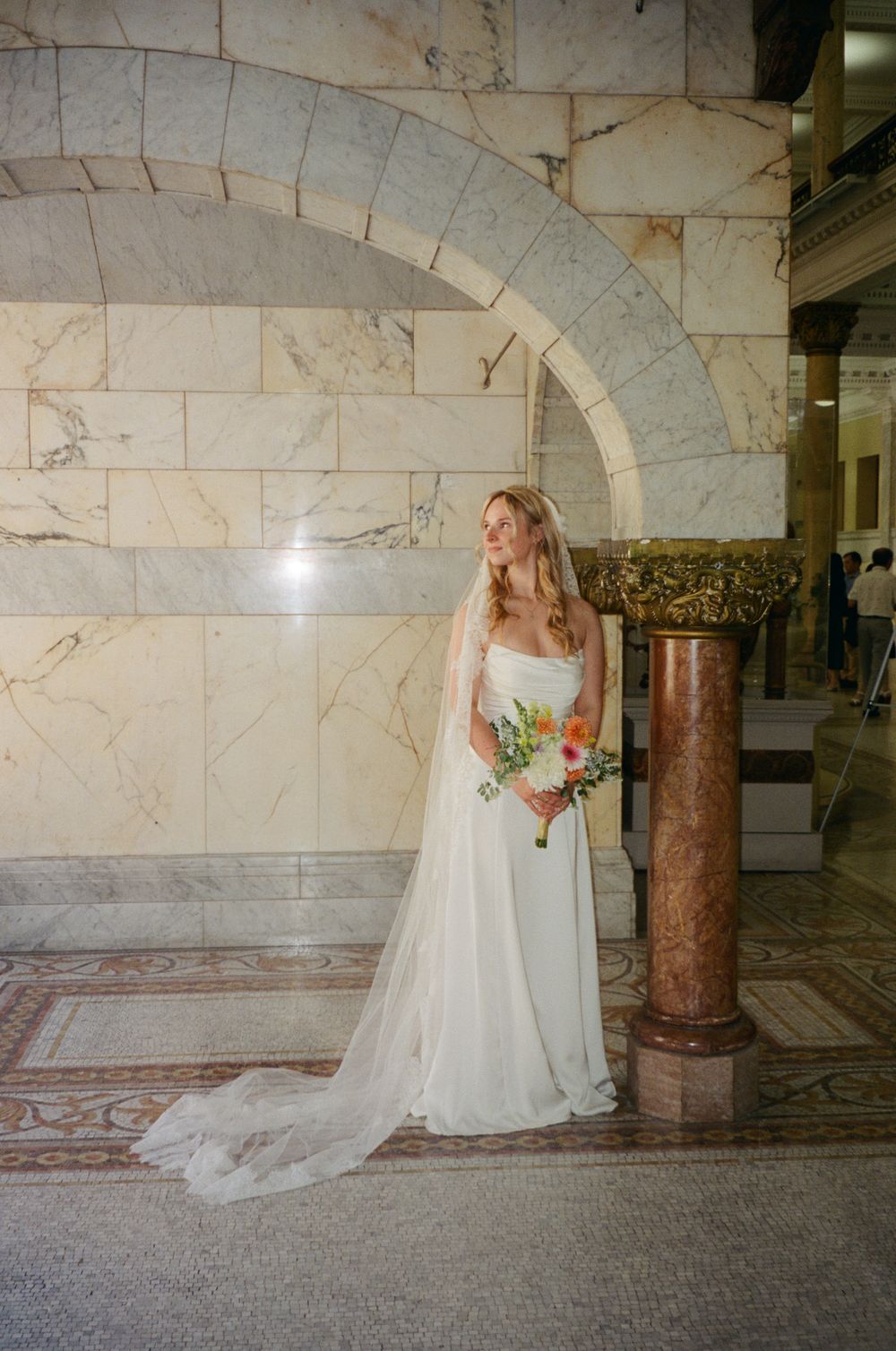 Bride in a flowing white gown and cathedral veil stands beside a marble column in an elegant hall.