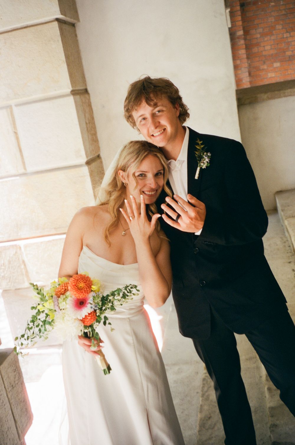 Happy newlyweds show off their wedding rings, bride in white gown holding a bright floral bouquet.