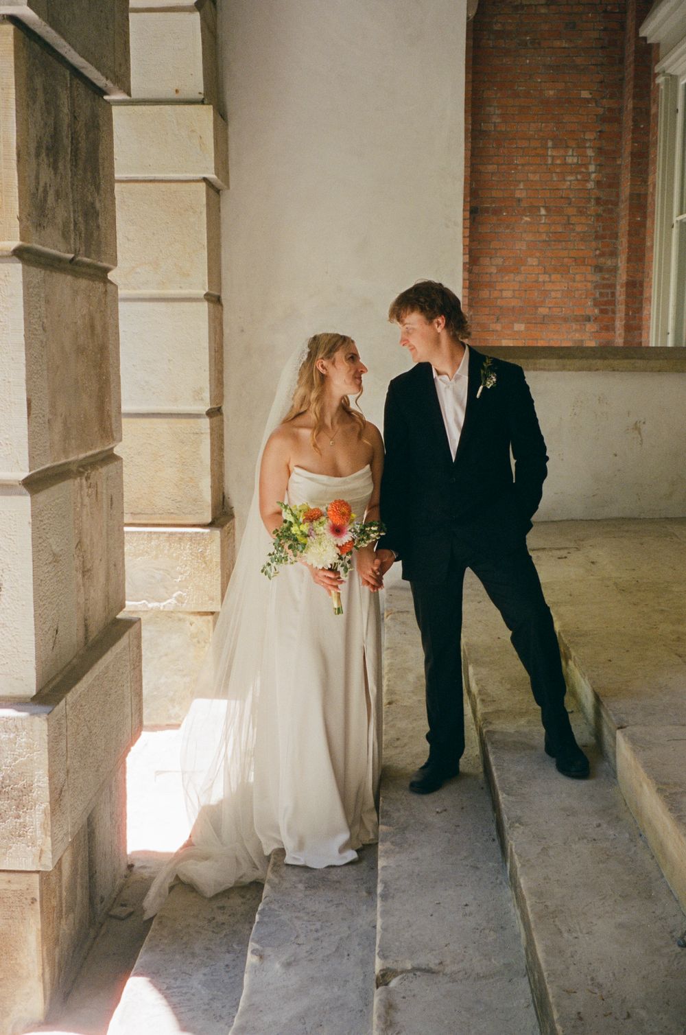 Bride and groom share a tender look on stone steps, bride in strapless gown holding a vibrant bouquet.