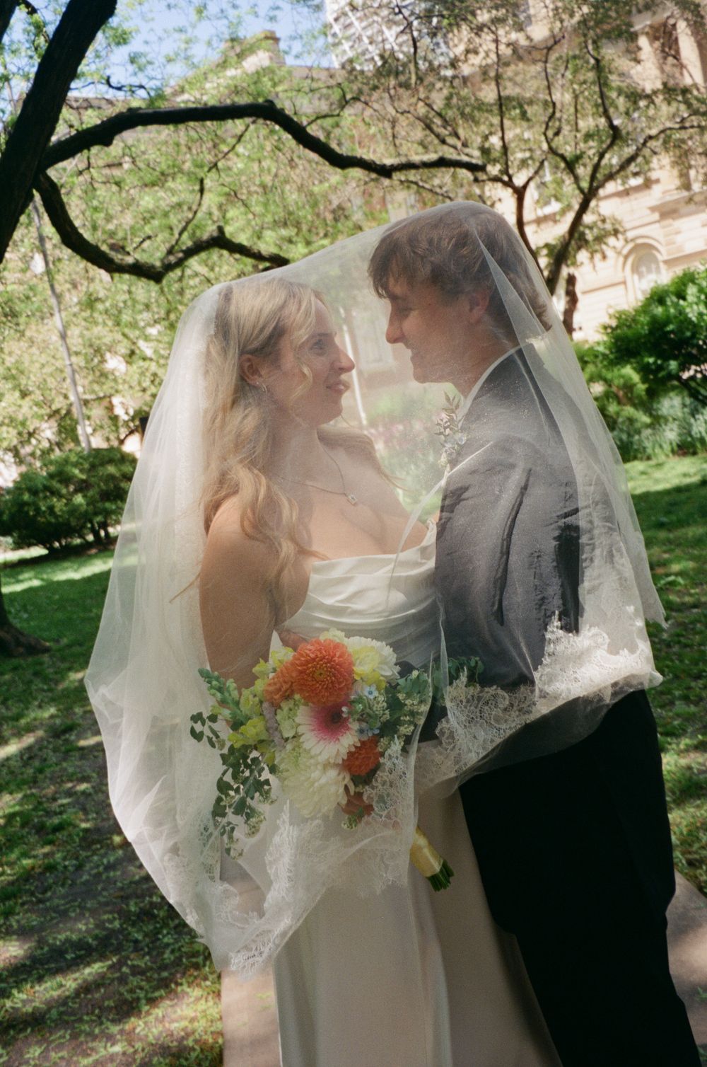 Bride and groom gaze at each other under a sheer lace veil in a sunlit garden setting.