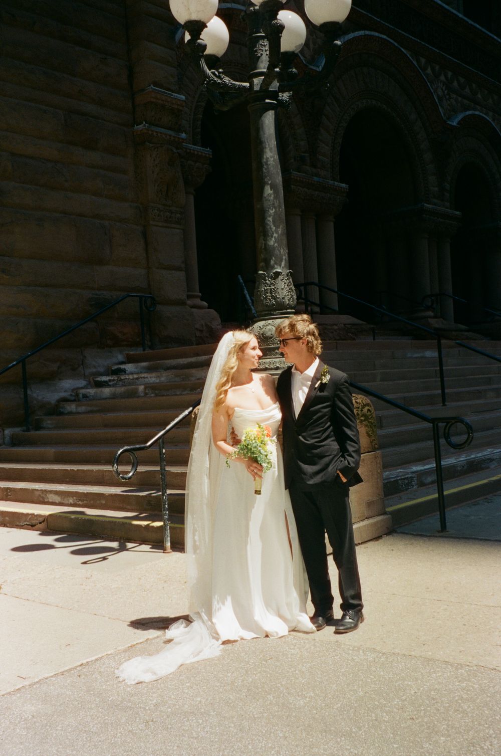 Bride and groom pose near an ornate lamppost outside a grand historic building on their wedding day.