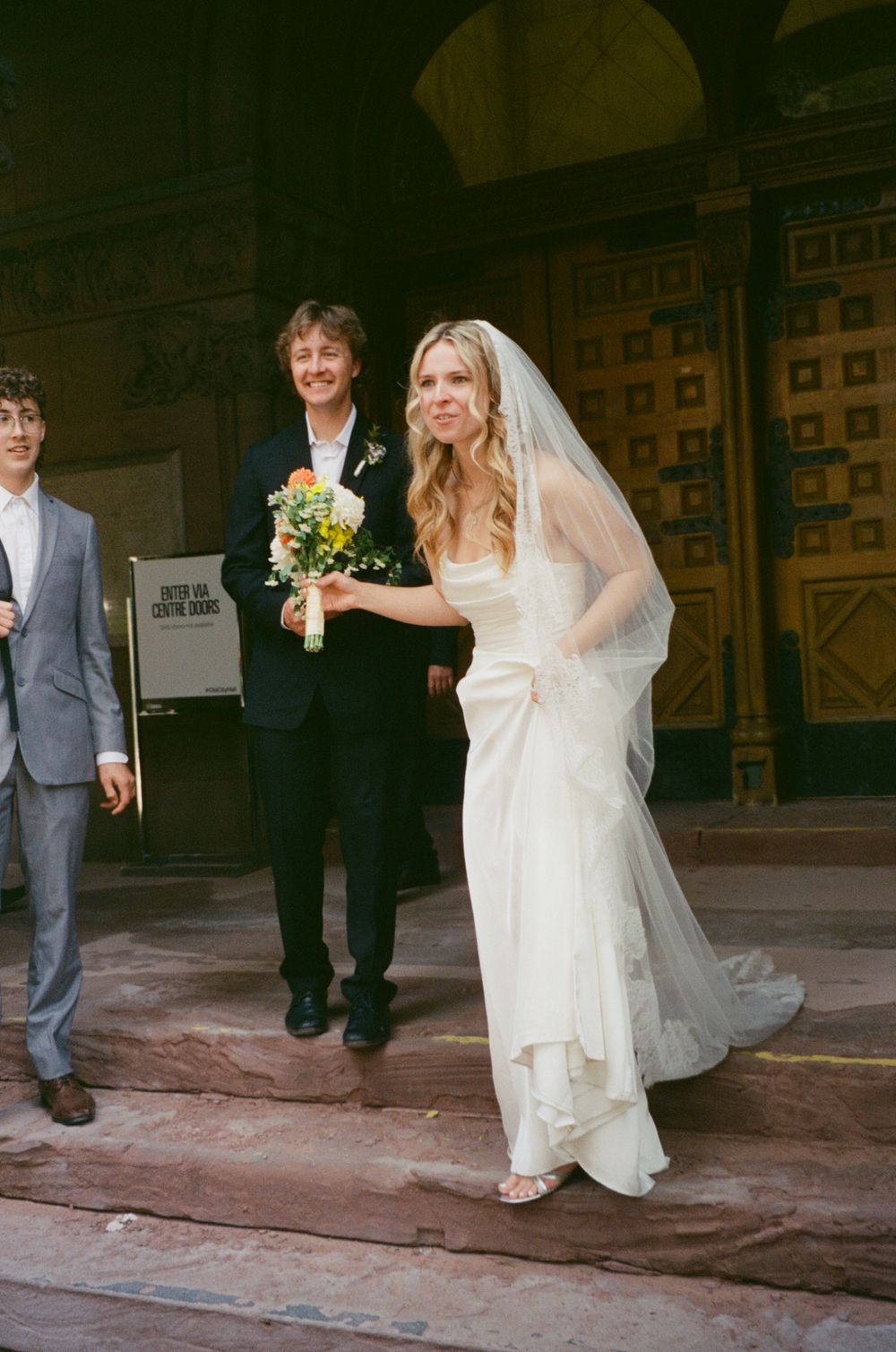 Smiling bride in white gown and veil exits a grand building with groom and guest beside her.