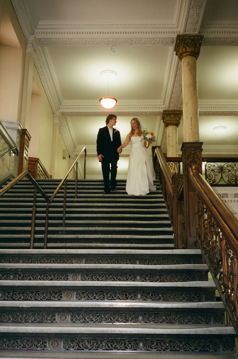 Bride and groom walk hand in hand down a grand ornate staircase inside a historic building.