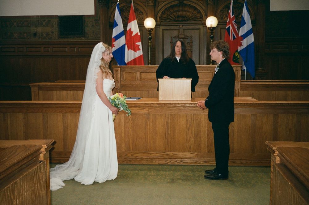 Bride and groom exchange vows before a judge in a wood-paneled courtroom with Canadian flags.
