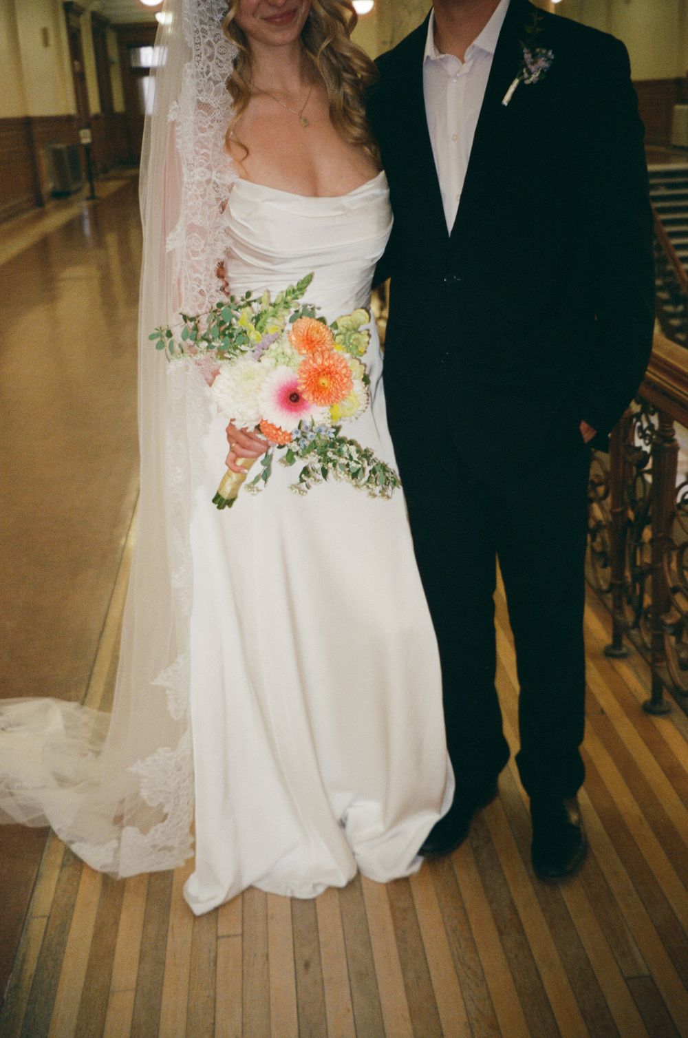 Bride in white gown with lace veil holds colorful bouquet beside groom in black suit.