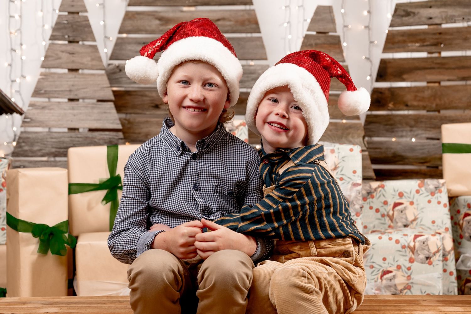 Two cheerful boys in Santa hats smile together surrounded by Christmas gifts in a festive holiday setting.