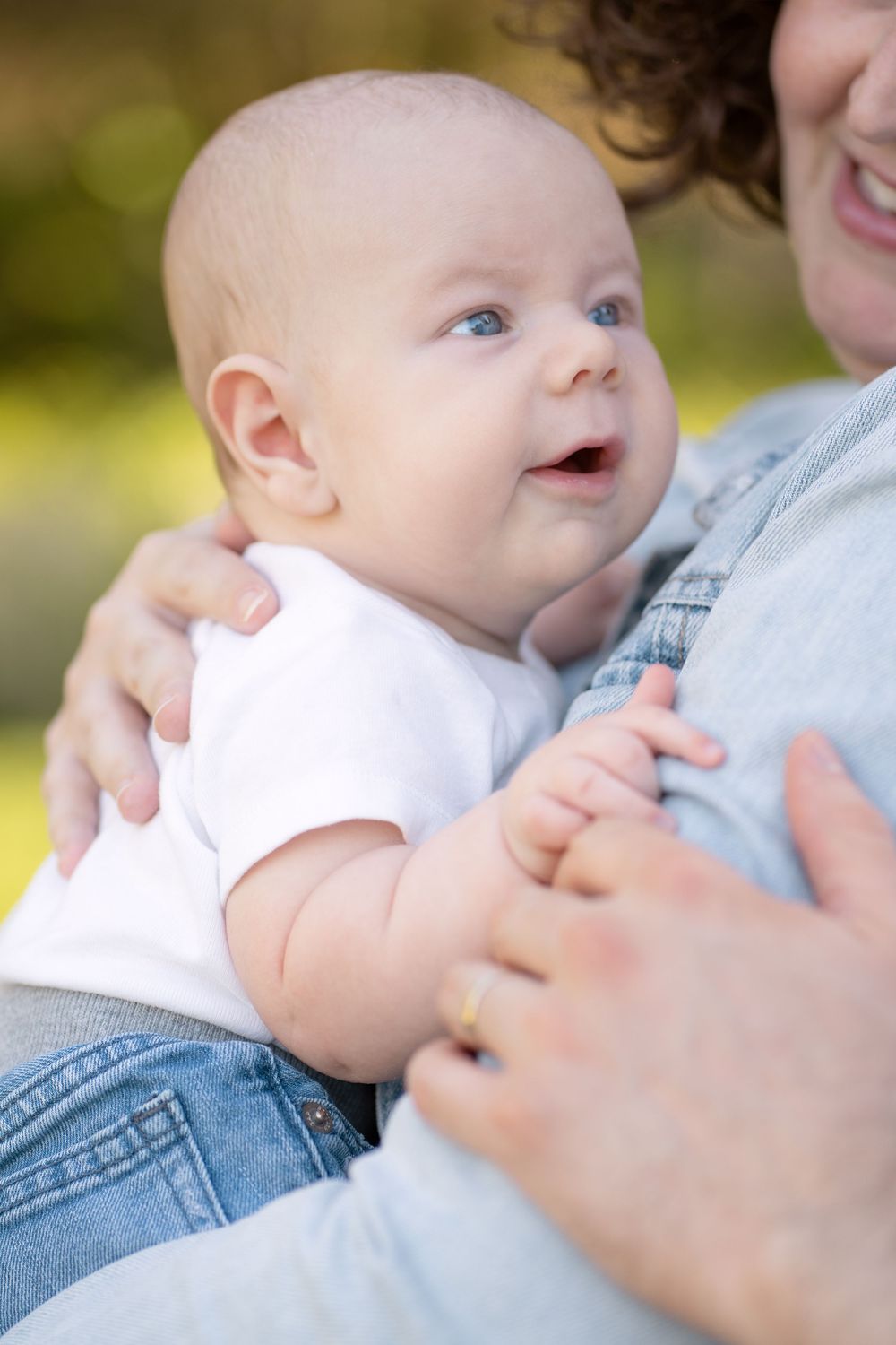 Adorable blue-eyed baby smiling while being held by mother wearing denim jacket outdoors in natural light.