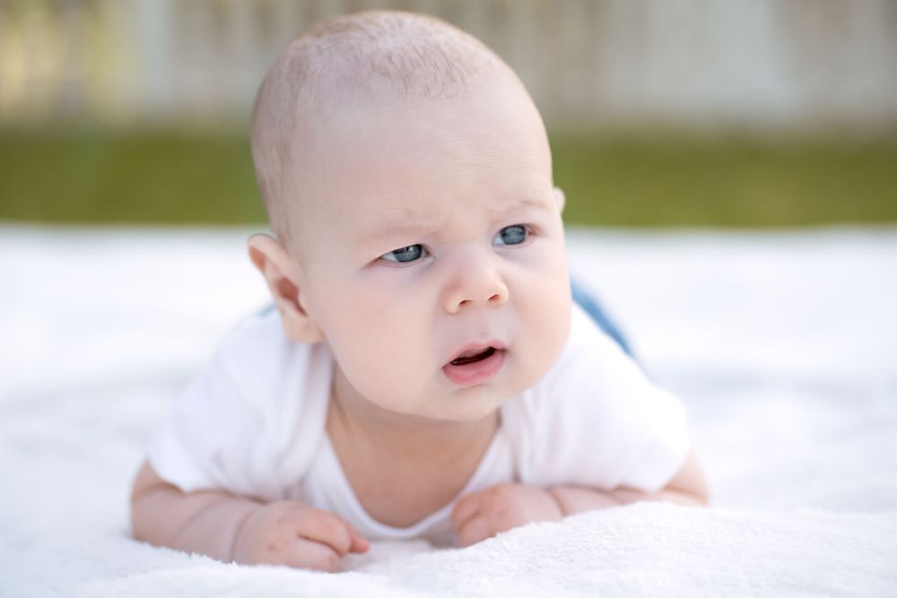 Cute baby in white onesie doing tummy time on a soft white blanket, looking up with bright blue eyes.