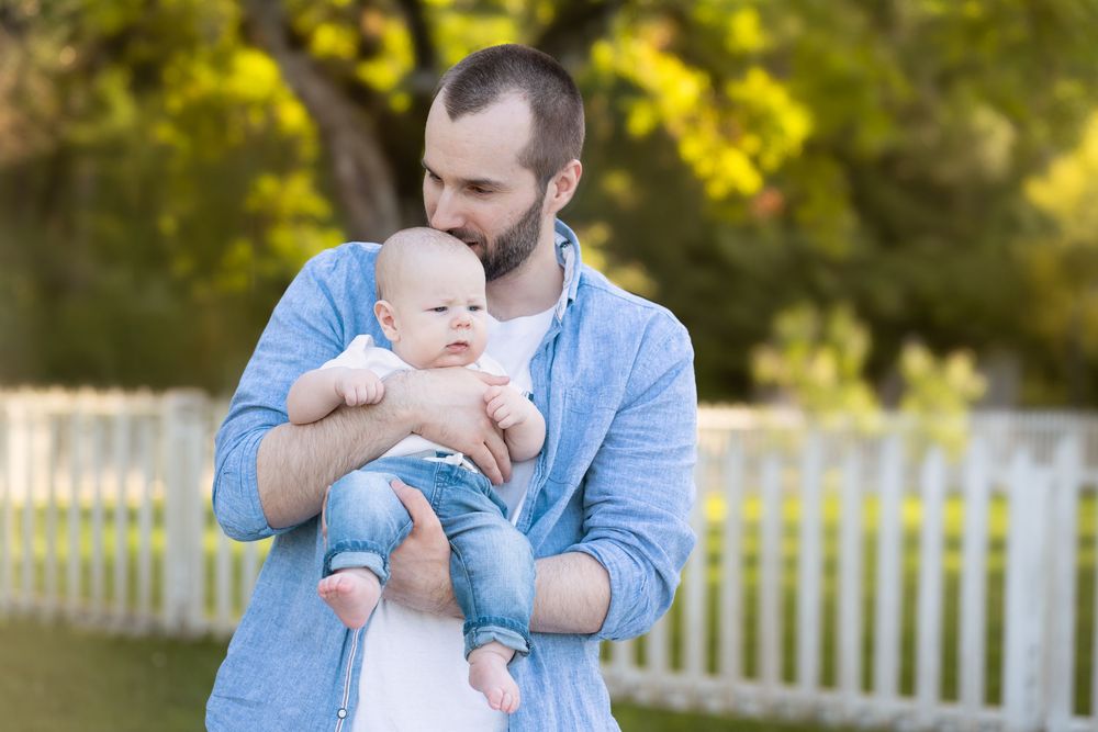 Father holding baby outdoors near white picket fence with green trees in background on sunny day.