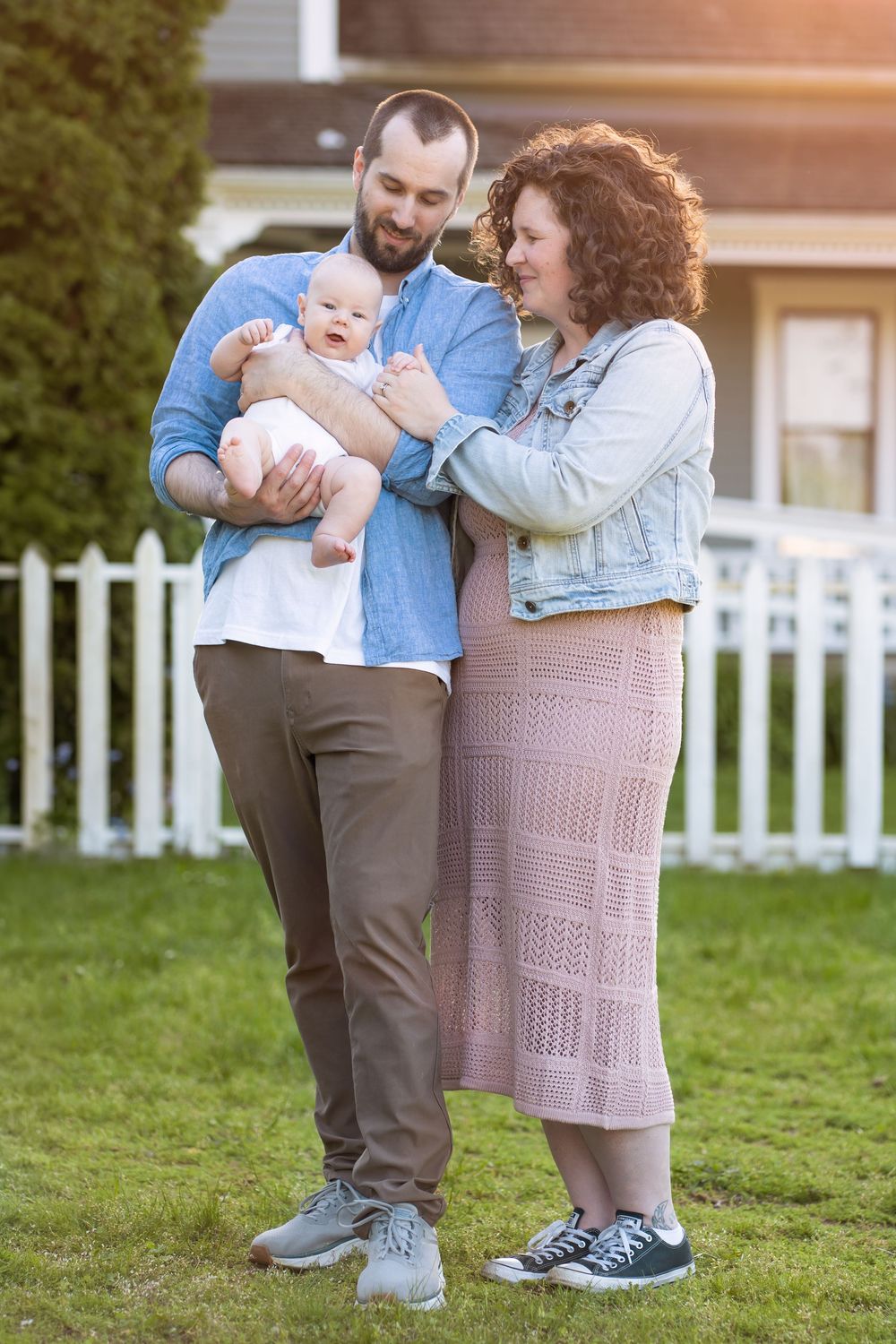 Young family with bearded dad, curly-haired mom in pink dress, holding newborn baby outdoors by white picket fence.