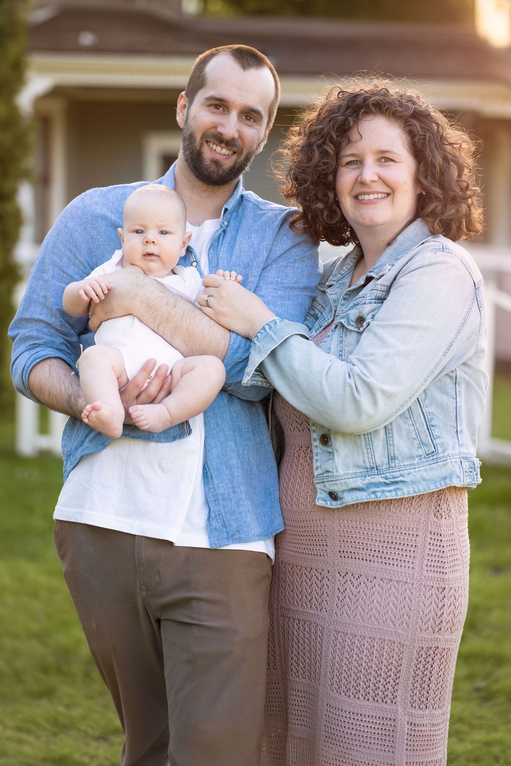 Happy young family with baby posing outdoors in front of a house on a sunny day.
