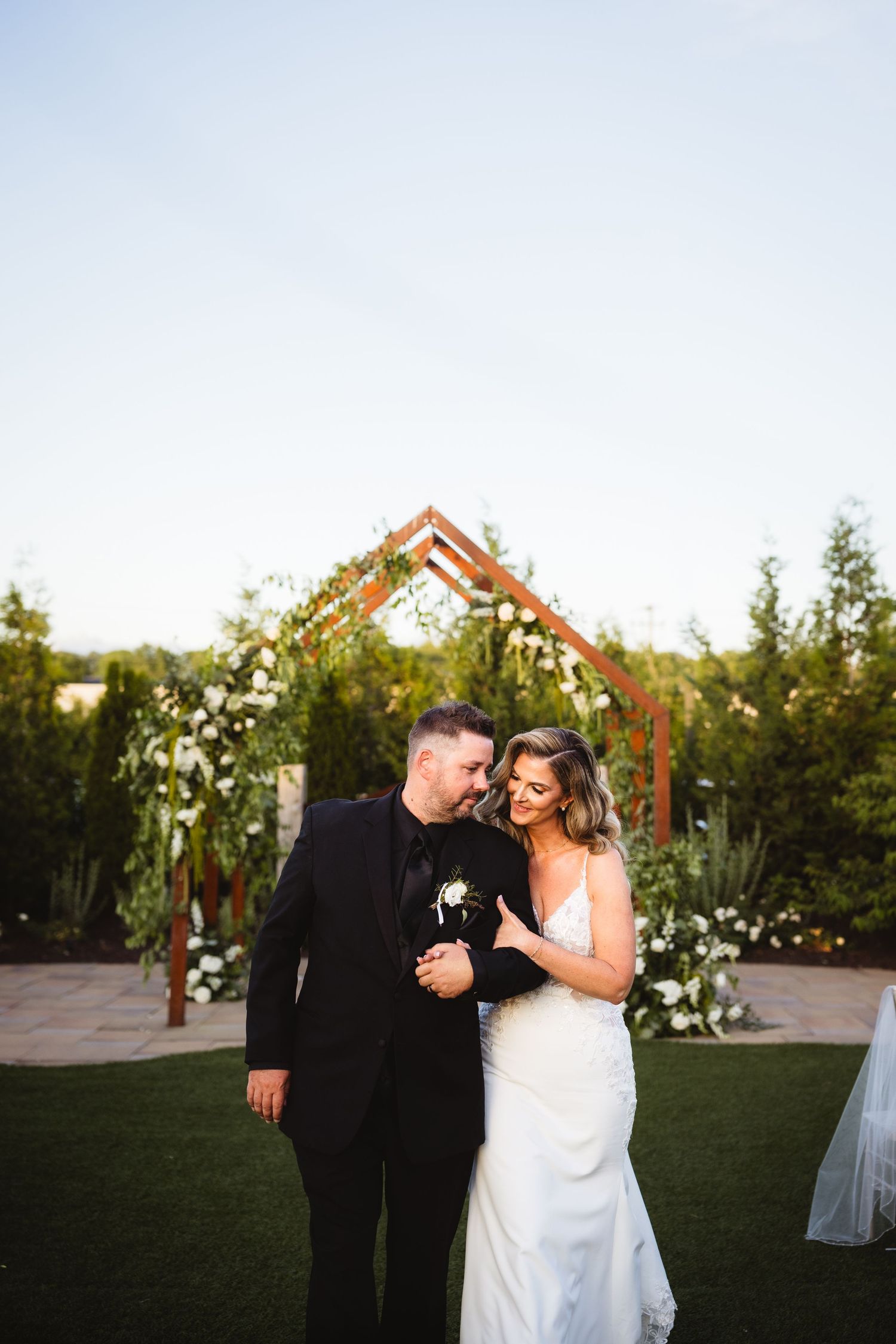 Bride and groom smiling together under a geometric wooden arch with white floral arrangements at an outdoor wedding ceremony.