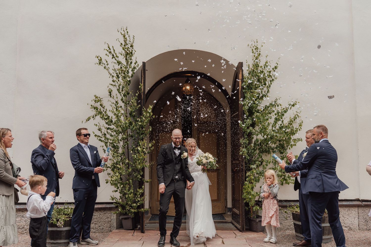 Newlyweds exit church doors as guests throw confetti during outdoor wedding ceremony.