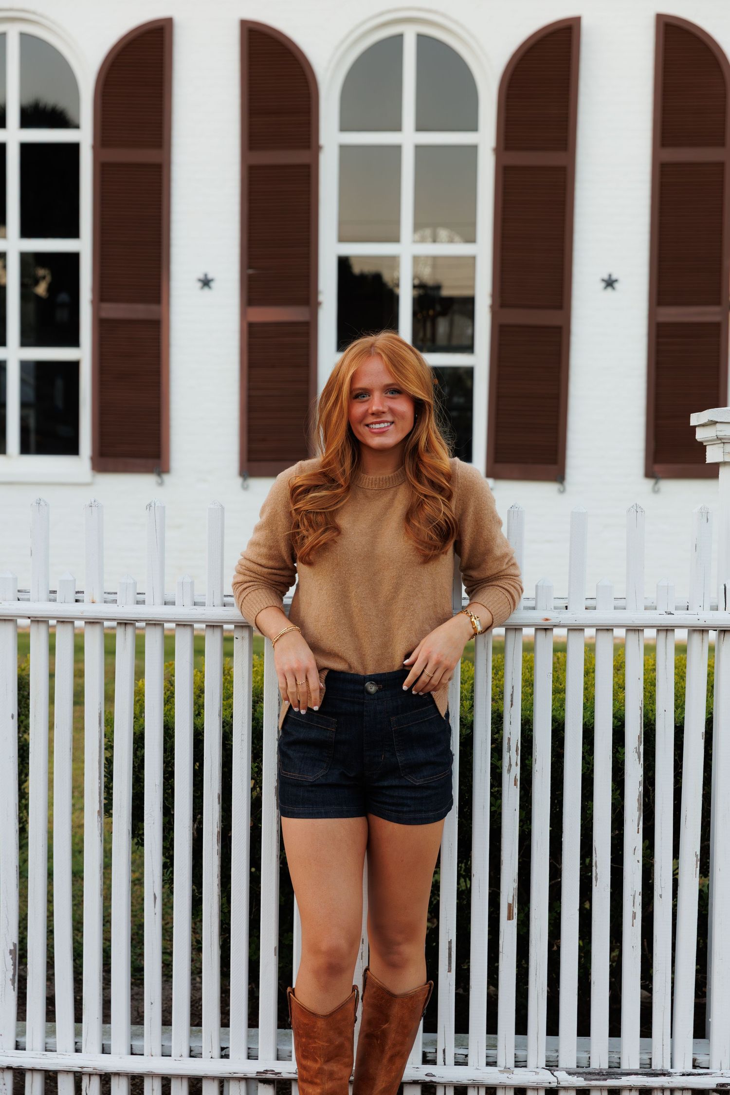 Young woman with red hair wearing a tan sweater, denim shorts, and brown boots stands by a white picket fence.