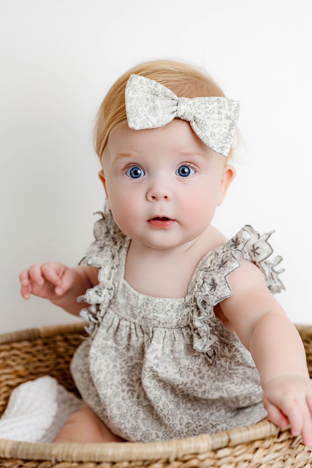 Adorable baby girl with blue eyes wearing a floral gray dress and matching bow headband sitting in a wicker basket.