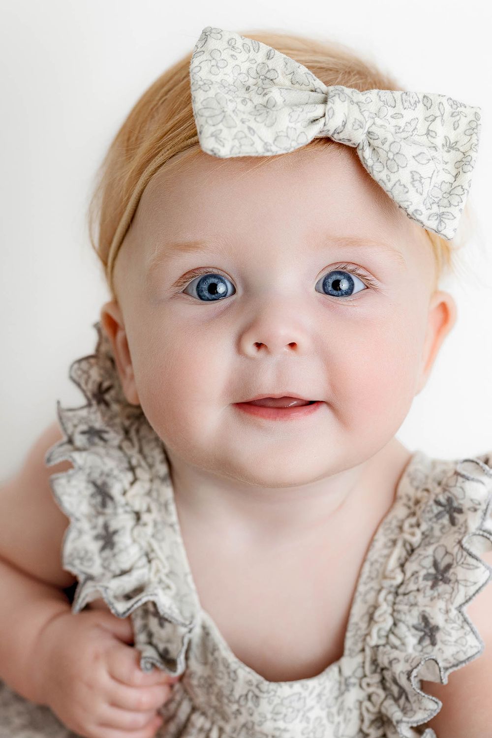 Adorable baby girl with blue eyes wearing a floral headband bow and ruffled dress, smiling at the camera.