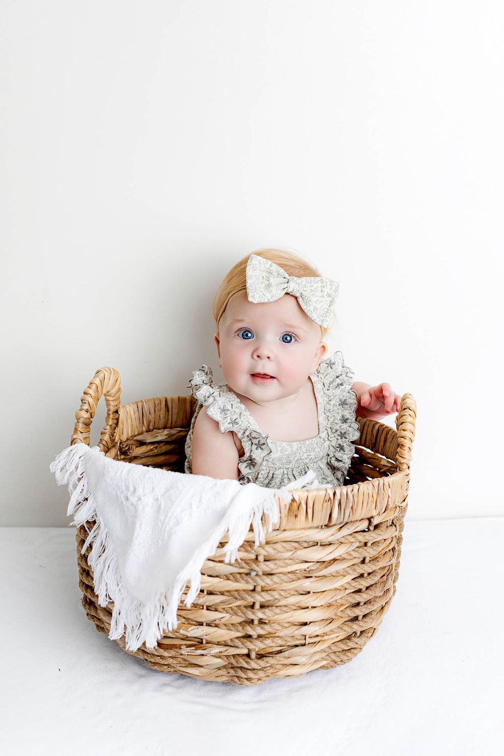 Adorable baby girl in gray ruffled romper and bow headband sitting in a wicker basket with white blanket.