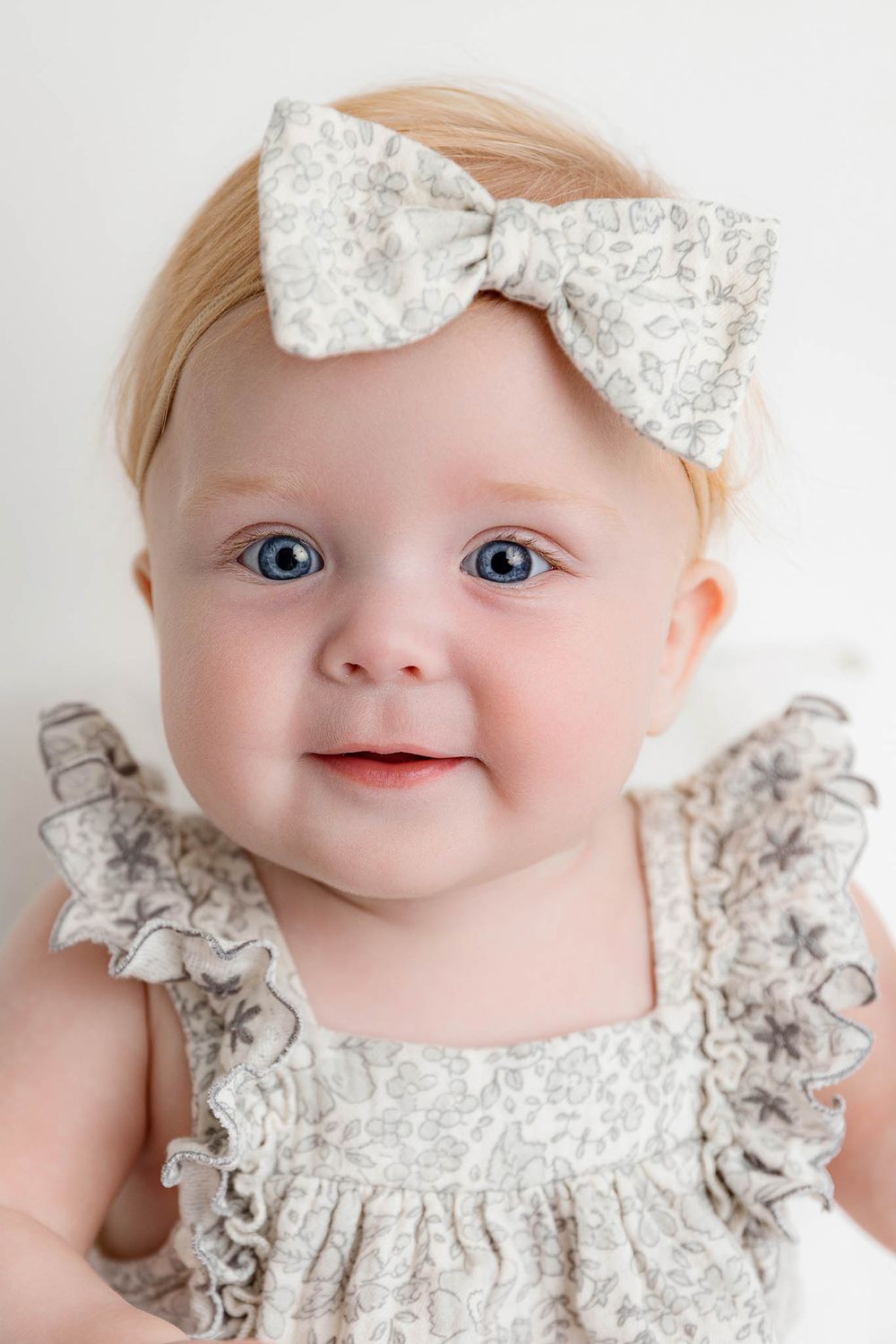 Adorable baby girl with blue eyes wearing a floral dress and matching bow headband, smiling sweetly.