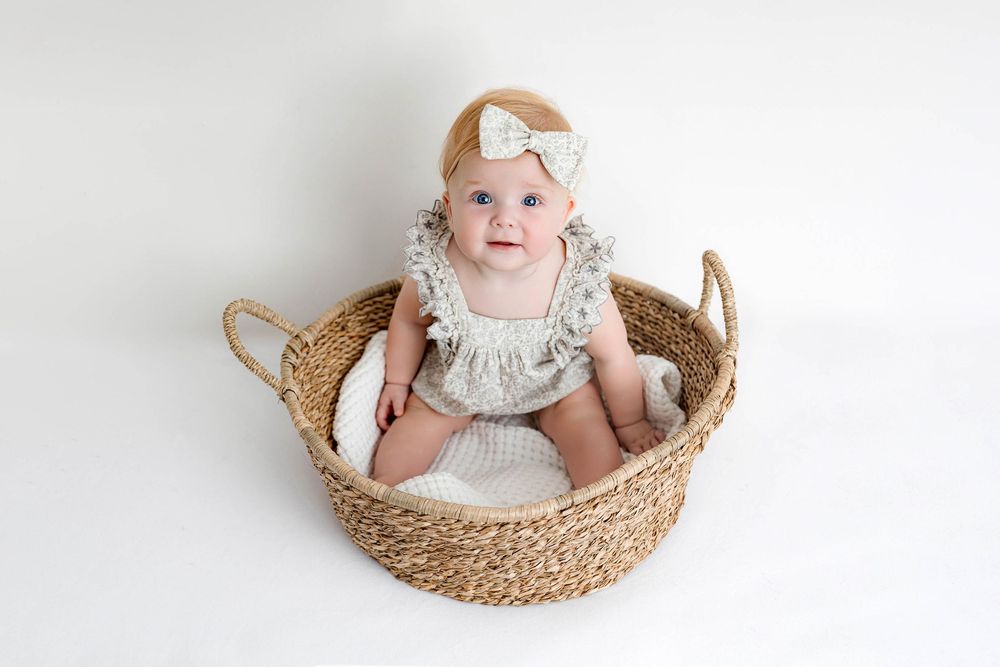 Adorable baby girl in a lace romper and bow headband sitting in a woven basket on a white background.